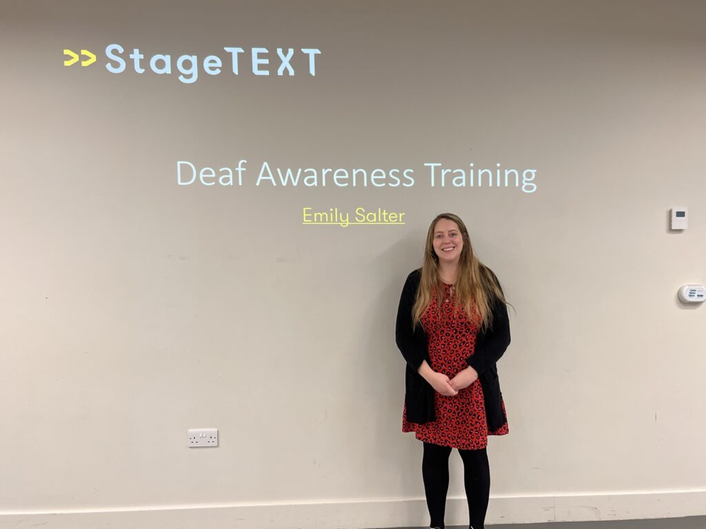 A projected screen onto a white wall saying stagetext, deaf awareness training Emily Salter. Standing in front is a white, blonde haired woman with a red dress and a black cardigan.