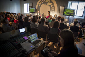 A Live subtitled event at the Wellcome Collection with a speaker in front of a room full of people and two screen either side full of text. At the forefront is the live subtitler on their machine typing what is being said live onto the screens.